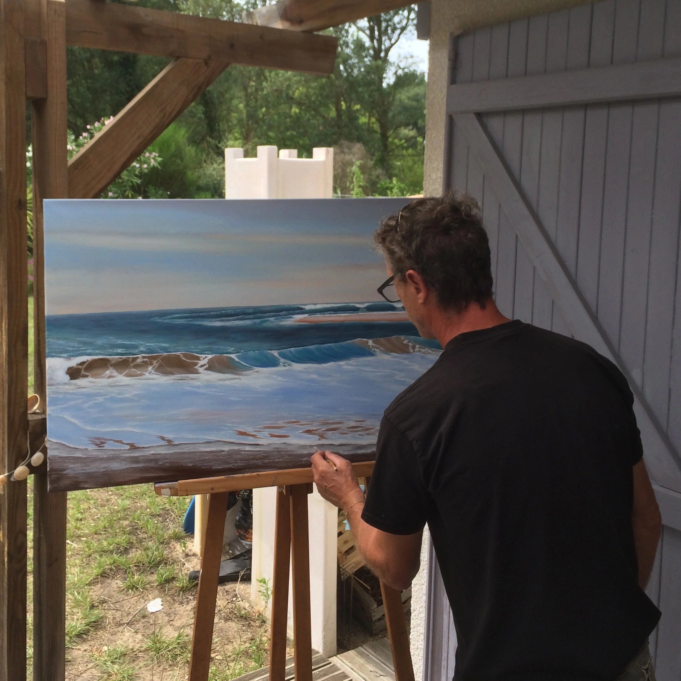 L’artiste Franck Costes en train de peindre une scène marine réaliste sur toile en extérieur — peinture à l’huile représentant une vague sur la plage.